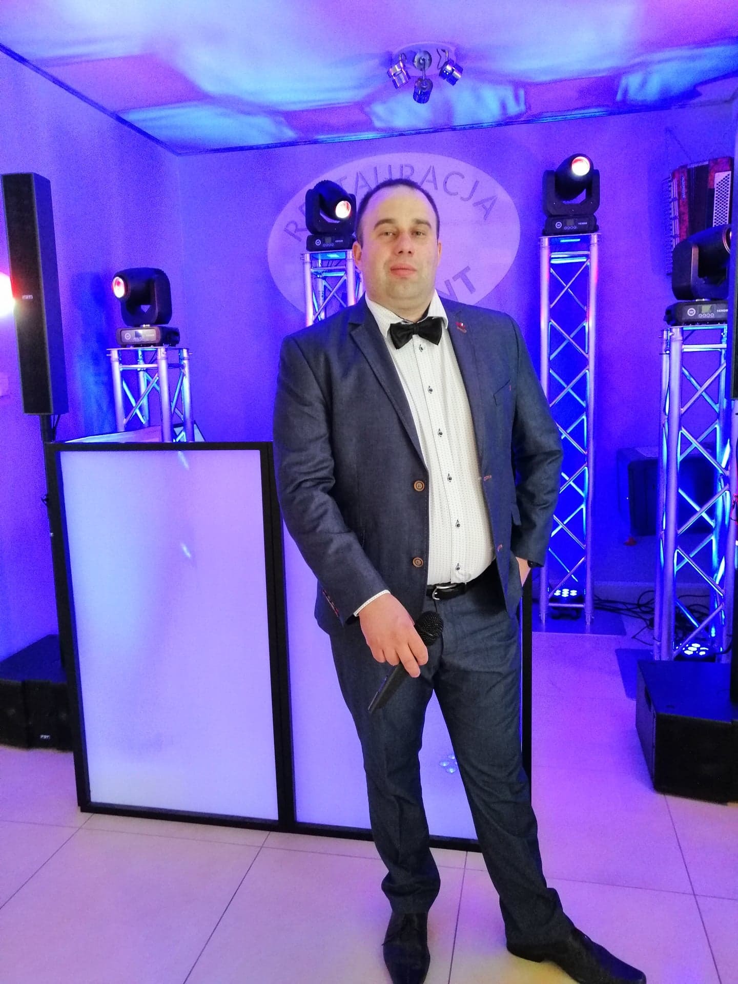 Mężczyzna w garniturze z mikrofonem na tle oświetlenia scenicznego w odcieniach fioletu i niebieskiego, widoczny DJ booth i kolumny głośnikowe.