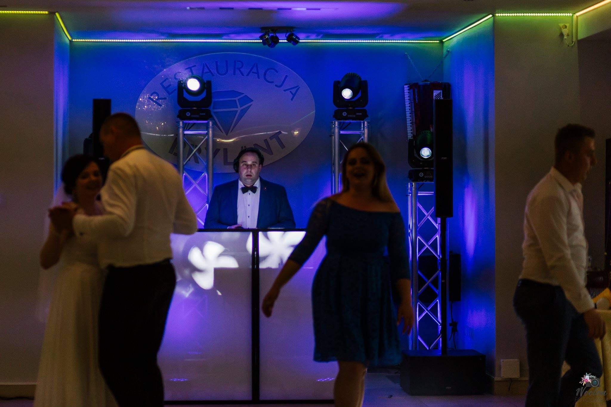 Dj w muszce za konsoletą z napisem 'Restauracja Diament', oświetlenie sceniczne i tańczący goście weselni w tle.