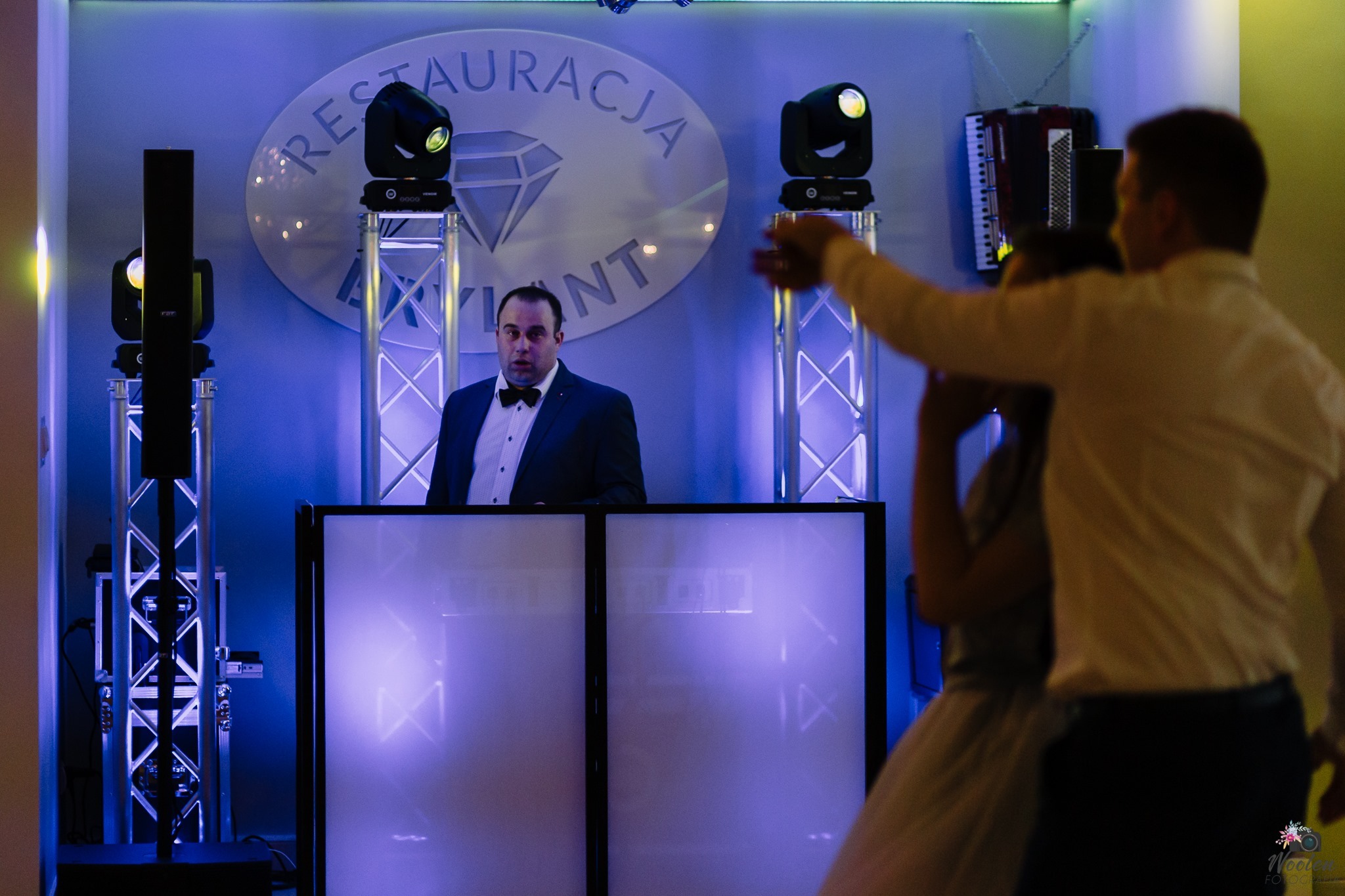DJ w eleganckim stroju za konsoletą, oświetlenie sceniczne i dekoracja 'Restauracja Brylant' w tle, para tańcząca w pierwszym planie.