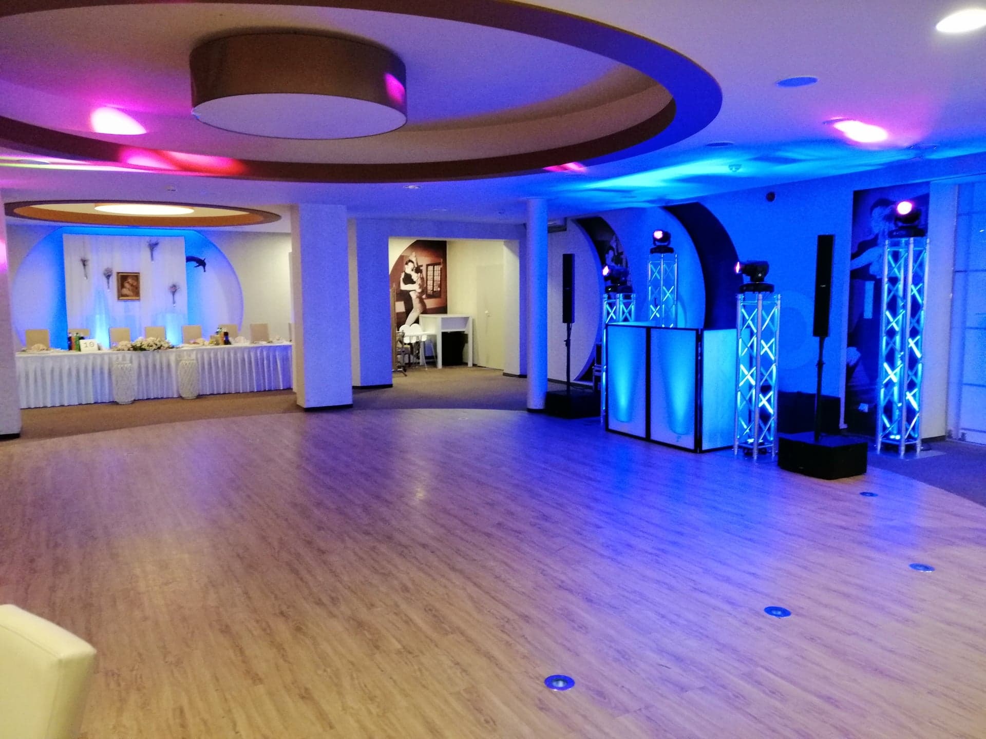 Sala bankietowa z oświetleniem w kolorze niebieskim i różowym, przygotowana na imprezę, widoczny stół z dekoracjami i sprzęt nagłośnieniowy.
