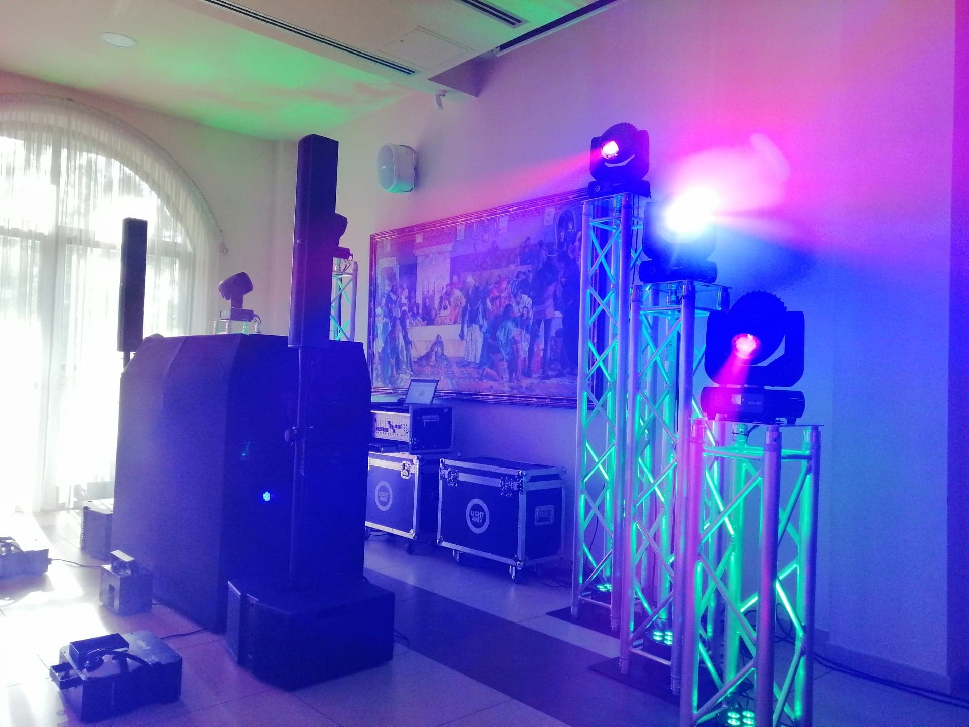 Profesjonalne nagłośnienie i oświetlenie sceniczne z reflektorami na kratownicach, kolumnami głośnikowymi i skrzyniami transportowymi w jasnym pomieszczeniu z obrazem na ścianie.