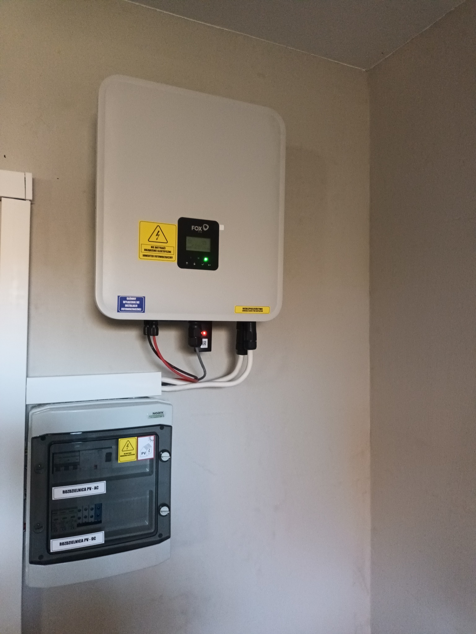 Zamontowany inwerter fotowoltaiczny marki FOX oraz rozdzielnica PV-AC/DC na ścianie wewnątrz pomieszczenia, widoczne przewody i naklejki ostrzegawcze.