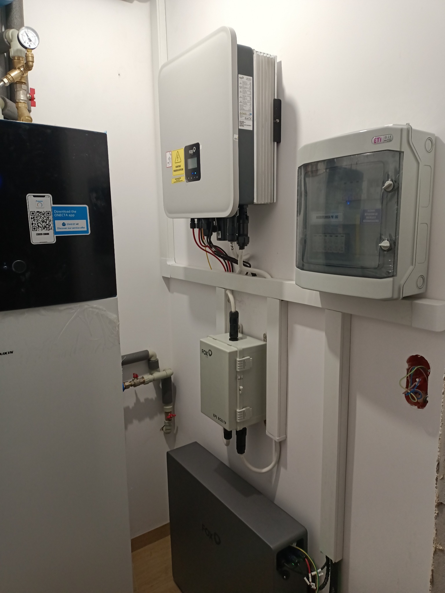 Zamontowany inwerter fotowoltaiczny FoxEss, skrzynka ETI, magazyn energii FoxEss oraz pompa ciepła Daikin z naklejką aplikacji ONECTA na białej ścianie wewnątrz budynku.