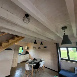 Jasne wnętrze drewnianego domu w Leśnej z widoczną jadalnią, schodami i oknem na zewnątrz, utrzymane w minimalistycznym stylu.