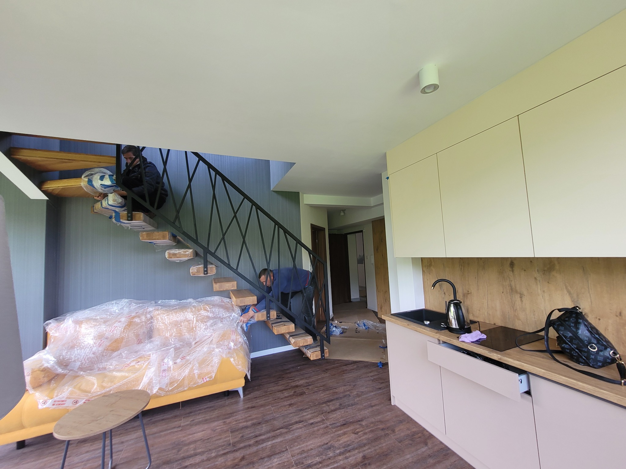 Dwaj mężczyźni montują drewniane stopnie schodów w nowoczesnym wnętrzu mieszkania; sofa zabezpieczona folią ochronną, widoczne kartony i elementy wyposażenia.