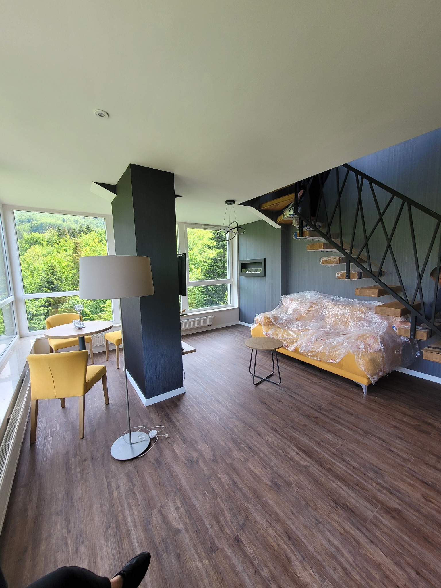 Wnętrze mieszkania po remoncie: żółta sofa zabezpieczona folią, schody z drewnianymi stopniami, widok na zielony las za oknem, stolik i krzesła w strefie jadalnej, brązowa podłoga