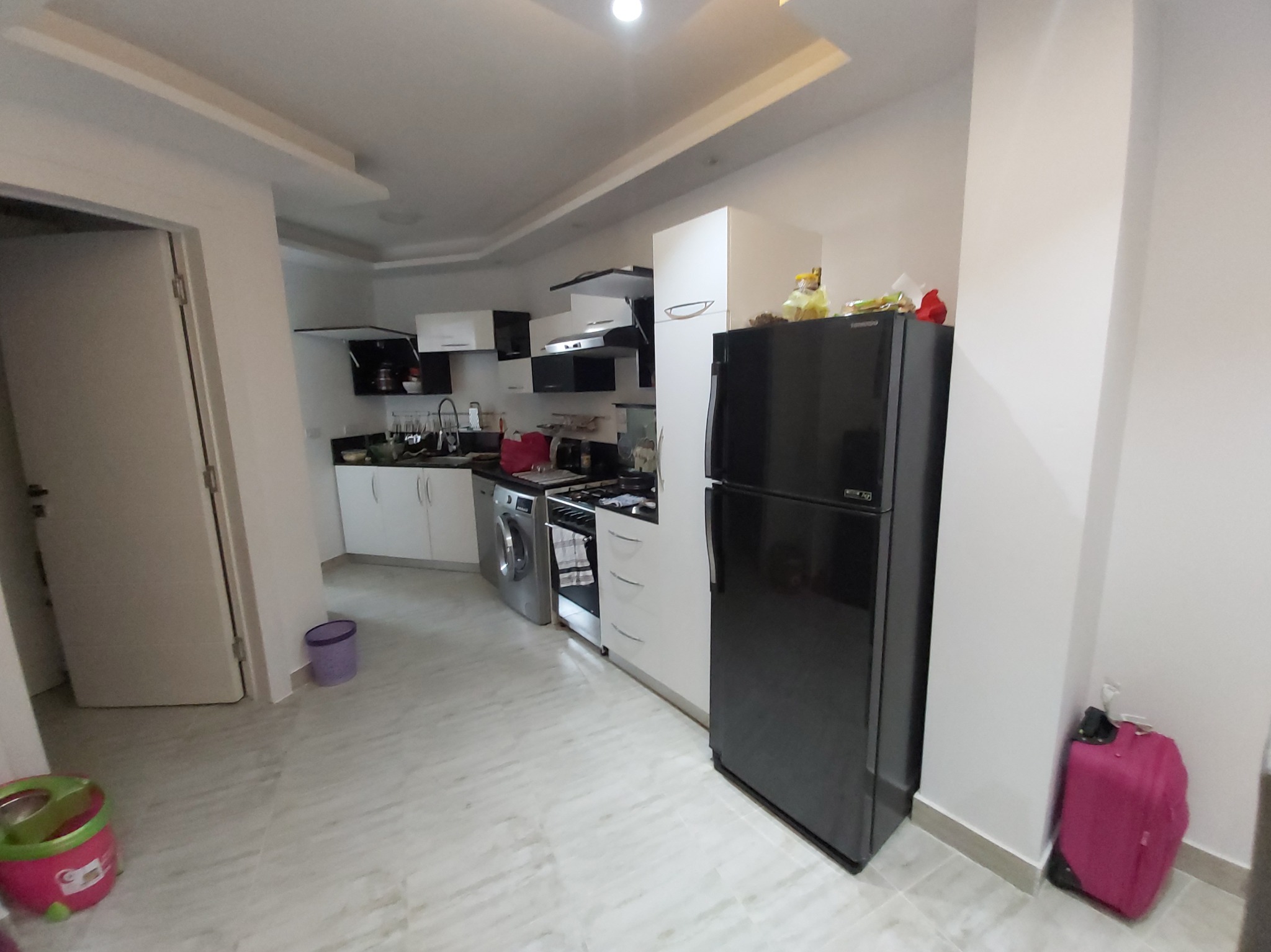 Ujęcie wnętrza kuchni w mieszkaniu z widoczną pralką, lodówką i kuchenką, z fioletowym wiaderkiem i różową walizką w tle.