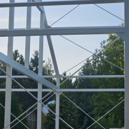 OFF Design Magdalena Domagalska - Biała, stalowa konstrukcja hali w trakcie budowy, widok przez szkielet na zieleń drzew i fragment budynku w tle, jasne niebo.