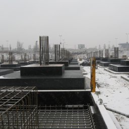 OFF Design Magdalena Domagalska - Zimowy krajobraz placu budowy z widocznymi fundamentami i zbrojeniami, pokrytymi śniegiem. Szara, pochmurna pogoda. W tle widoczne budynki i latarnie.