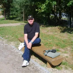 Mężczyzna w czarnej koszulce i jeansach siedzi na drewnianej ławce w parku, obok leży poduszka i torba.