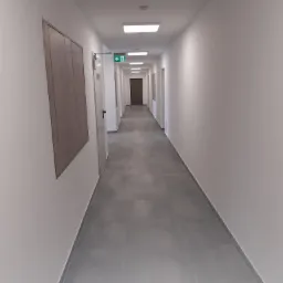 Długi, jasny korytarz z szarymi płytkami podłogowymi, białymi ścianami i drewnianymi drzwiami po lewej stronie, zakończony ciemnymi drzwiami w oddali, oświetlony przez równomiernie rozmieszczone...