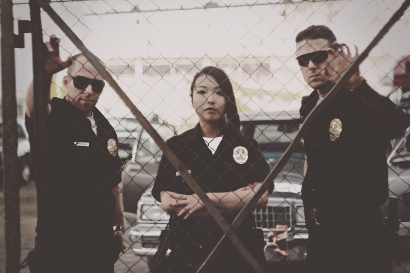 Trzech aktorów w stylizowanych mundurach policyjnych pozuje za metalową siatką na tle samochodów; produkcja filmowa.