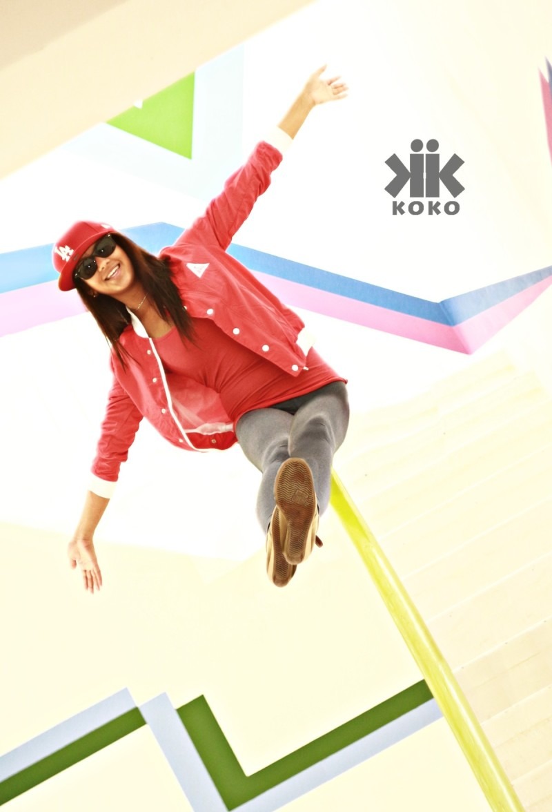Kobieta w czerwonej kurtce i czapce z daszkiem, z uniesionymi rękami, pozująca na tle geometrycznej ściany z logo 'Kik Koko'.