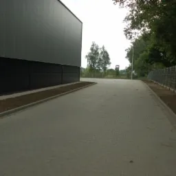 Ułożona szara kostka brukowa przed budynkiem przemysłowym z szarej blachy trapezowej i ogrodzeniem panelowym z siatki z widocznym fragmentem zieleni w tle.