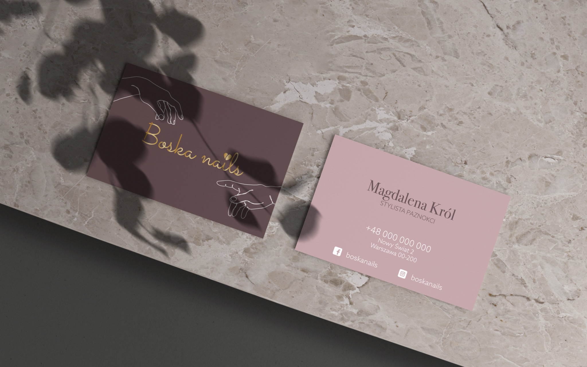 Dwie wizytówki na marmurowym tle w cieniu liści. Jedna w kolorze bordowym z napisem 'Boska nails' i rysunkiem dłoni, druga w kolorze pudrowego różu z danymi kontaktowymi stylistki paznokci...