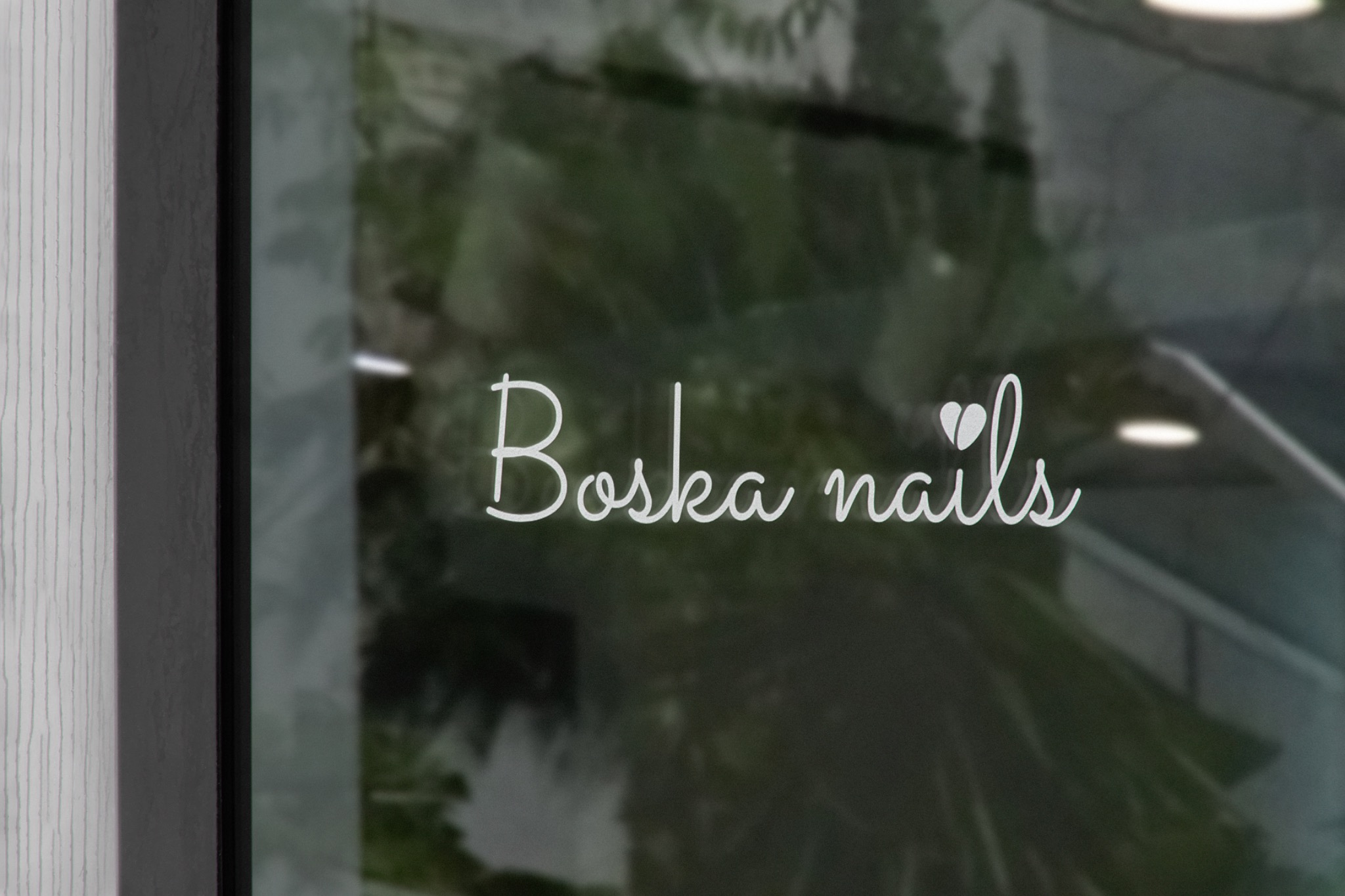 Elegancki napis 'Boska nails' w kolorze białym, umieszczony na szklanej witrynie salonu, z widocznym odbiciem zieleni w tle.
