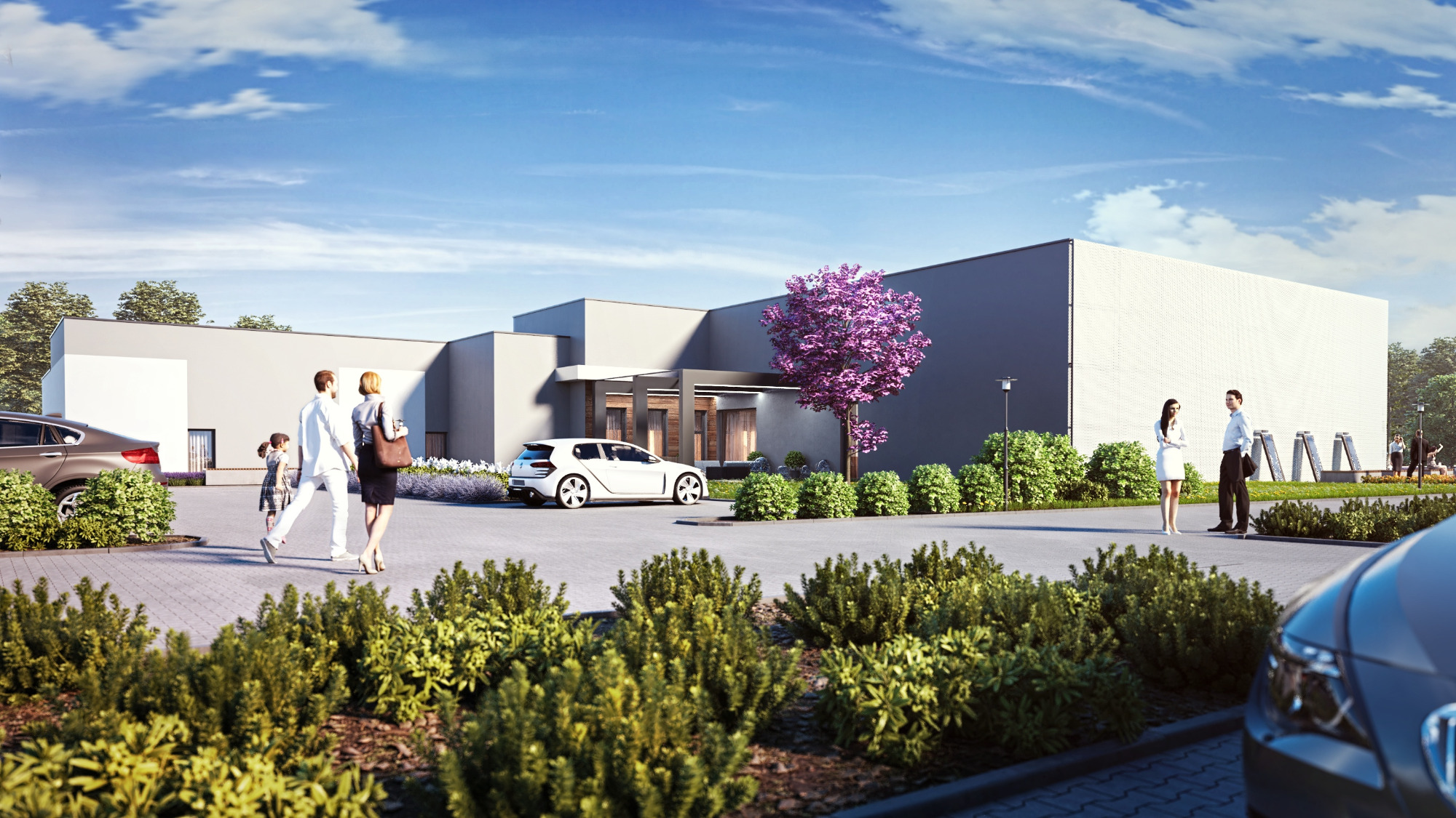 Nowoczesny budynek z szarej cegły z białymi akcentami, otoczony zielenią i drzewem o purpurowych kwiatach, z zaparkowanymi samochodami i spacerującymi ludźmi na parkingu.