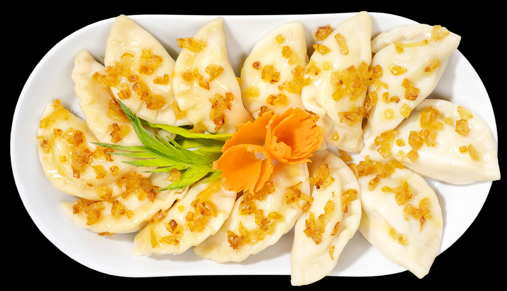 Porcja pierogów okraszonych zeszkloną cebulką, udekorowanych zielonymi gałązkami i marchewkowym kwiatkiem, podana na owalnym, białym talerzu na czarnym tle.