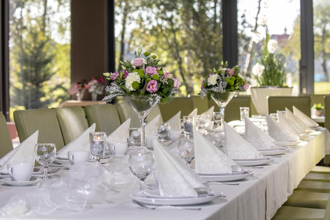 Elegancko nakryty stół z białym obrusem, zastawą, kieliszkami, serwetkami z koronkowym brzegiem i kompozycjami kwiatowymi w szklanych wazonach w kształcie kielichów martini, oświetlony naturalnym...