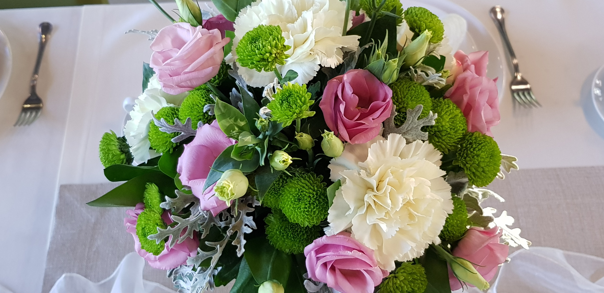 Dekoracja stołu z białych goździków, różowych róż i zielonych chryzantem kulistych, widelce po obu stronach, delikatna biała tkanina.