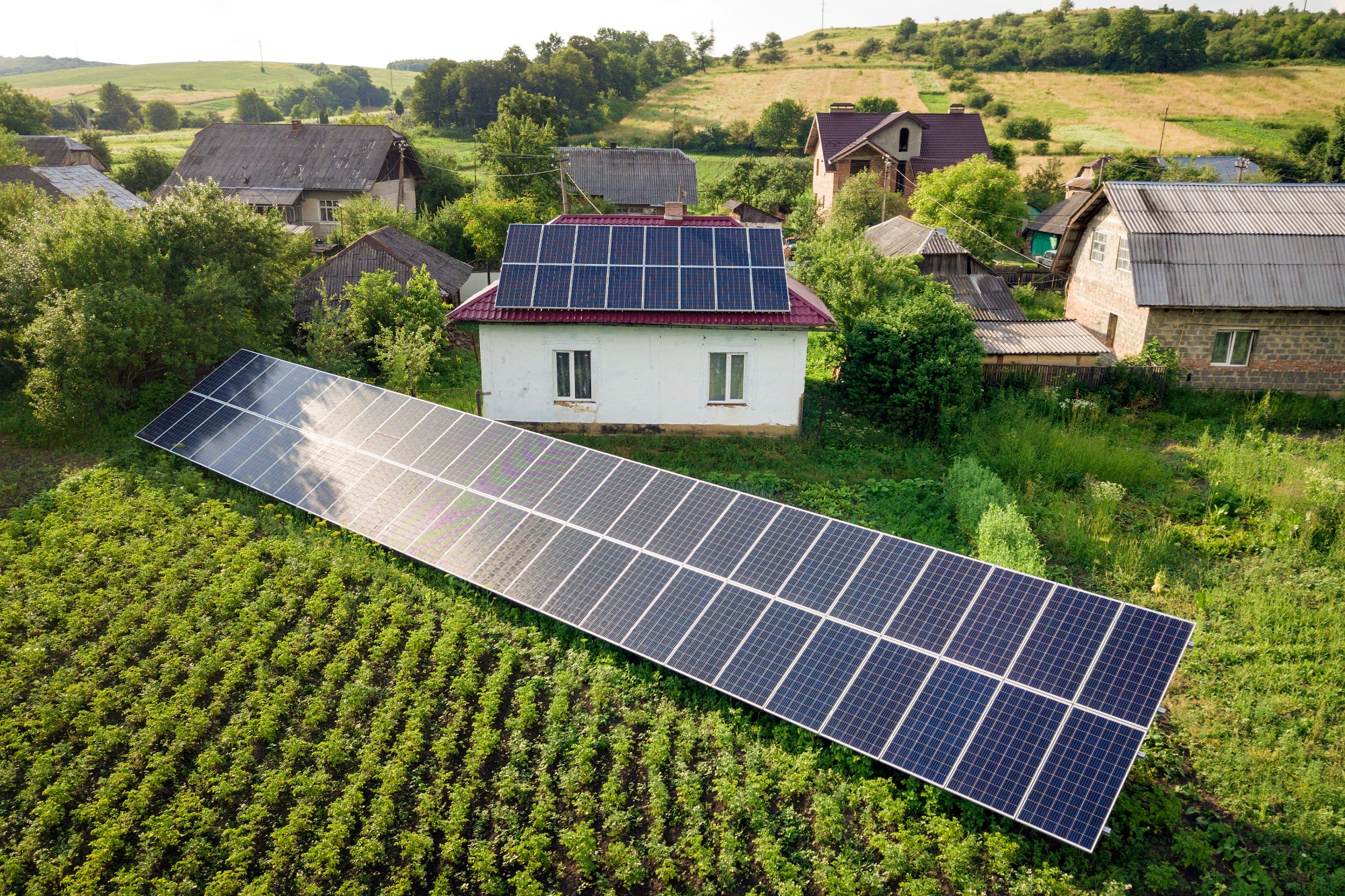 Rozległa instalacja fotowoltaiczna na gruncie obok domu z panelami na dachu, widok z góry na wiejski krajobraz.