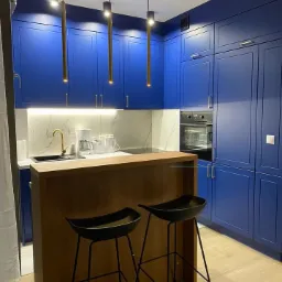 Niebieska kuchnia z drewnianym barkiem i dwoma czarnymi hokerami. Oświetlenie punktowe z trzema złotymi lampami. Marmurowy backsplash i sprzęt AGD w zabudowie.