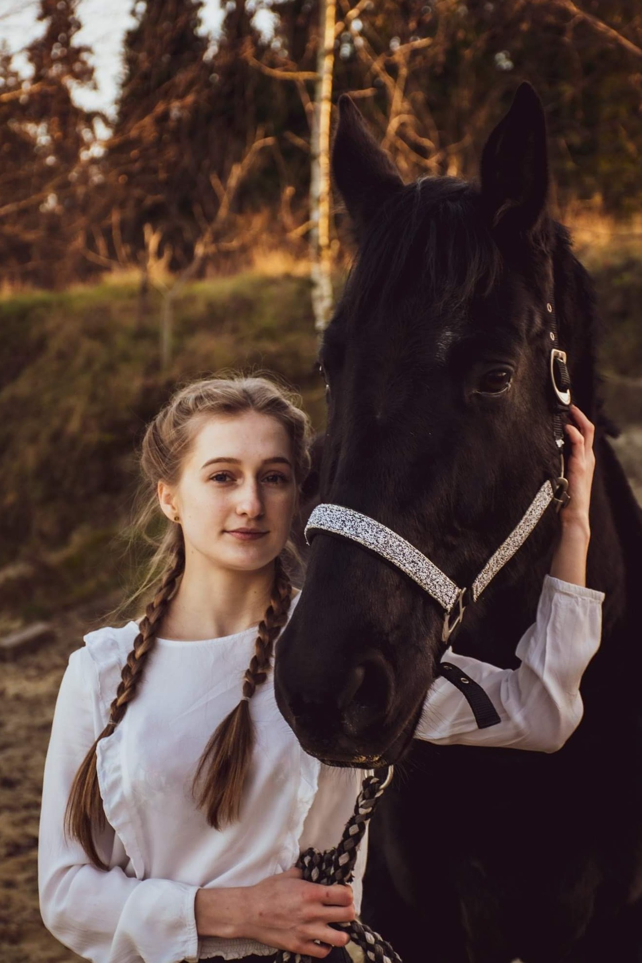 Portret młodej kobiety z blond warkoczami, w białej bluzce, stojącej obok czarnego konia z ozdobnym kantarem na tle leśnym.
