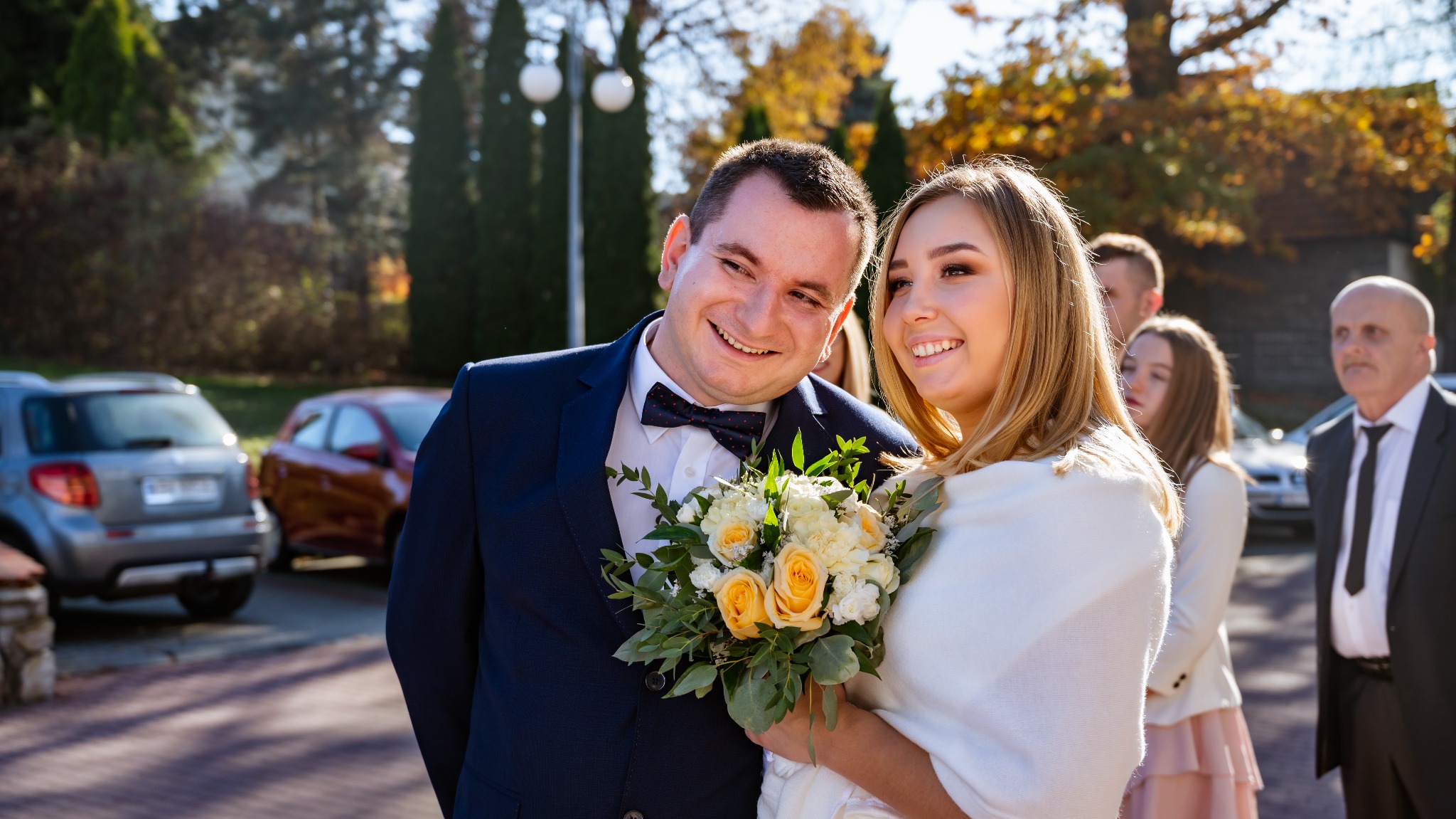 Uśmiechnięta para młoda w plenerze, on w granatowym garniturze z muchą w kropki, ona z bukietem żółtych i białych róż, w tle goście i samochody.