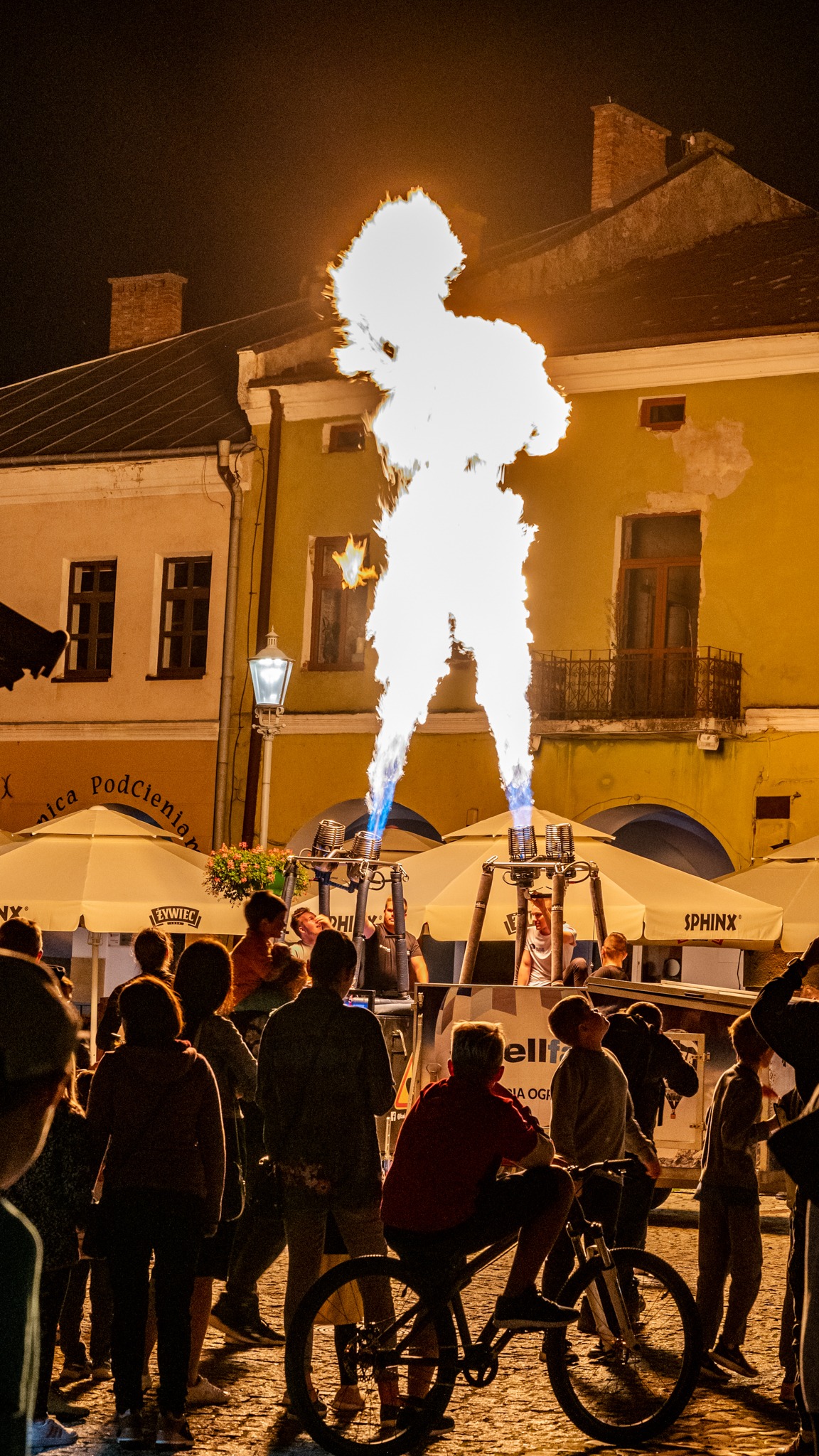 Nocny pokaz ognia z dwoma płomieniami skierowanymi ku górze, otoczony przez obserwujących ludzi na tle miejskiej architektury i parasoli kawiarnianych.