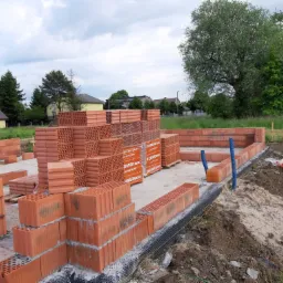 Wznoszenie ścian fundamentowych z czerwonej ceramiki budowlanej na przygotowanym podłożu, widoczne palety z materiałem i elementy izolacji pionowej.