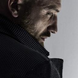 wilk fotografia - Profilowe ujęcie brodatego mężczyzny w ciemnej marynarce o fakturze w kratkę, z ostrym spojrzeniem w bok, oświetlonego z góry.