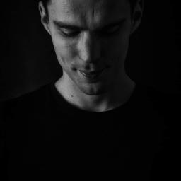 wilk fotografia - Czarno-biały portret młodego mężczyzny z lekkim uśmiechem, spoglądającego w dół, w ciemnym otoczeniu.