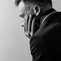 wilk fotografia - Czarno-białe zbliżenie profilu mężczyzny w garniturze, zamyślonego, z dłonią na twarzy.