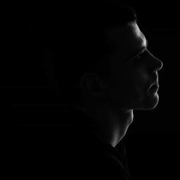 wilk fotografia - Czarno-biały portret profilowy mężczyzny, oświetlonego z boku, z wyraźnie zarysowanymi konturami twarzy na ciemnym tle.