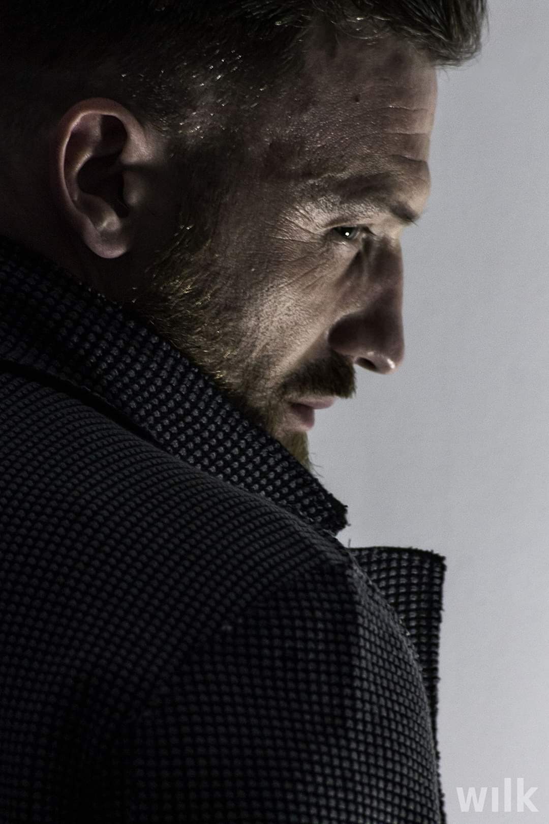 Profilowe ujęcie brodatego mężczyzny w ciemnej marynarce o fakturze w kratkę, z ostrym spojrzeniem w bok, oświetlonego z góry.