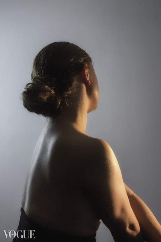 Portret od tyłu kobiety z upiętymi włosami, oświetlonej bocznym światłem, na szarym tle.