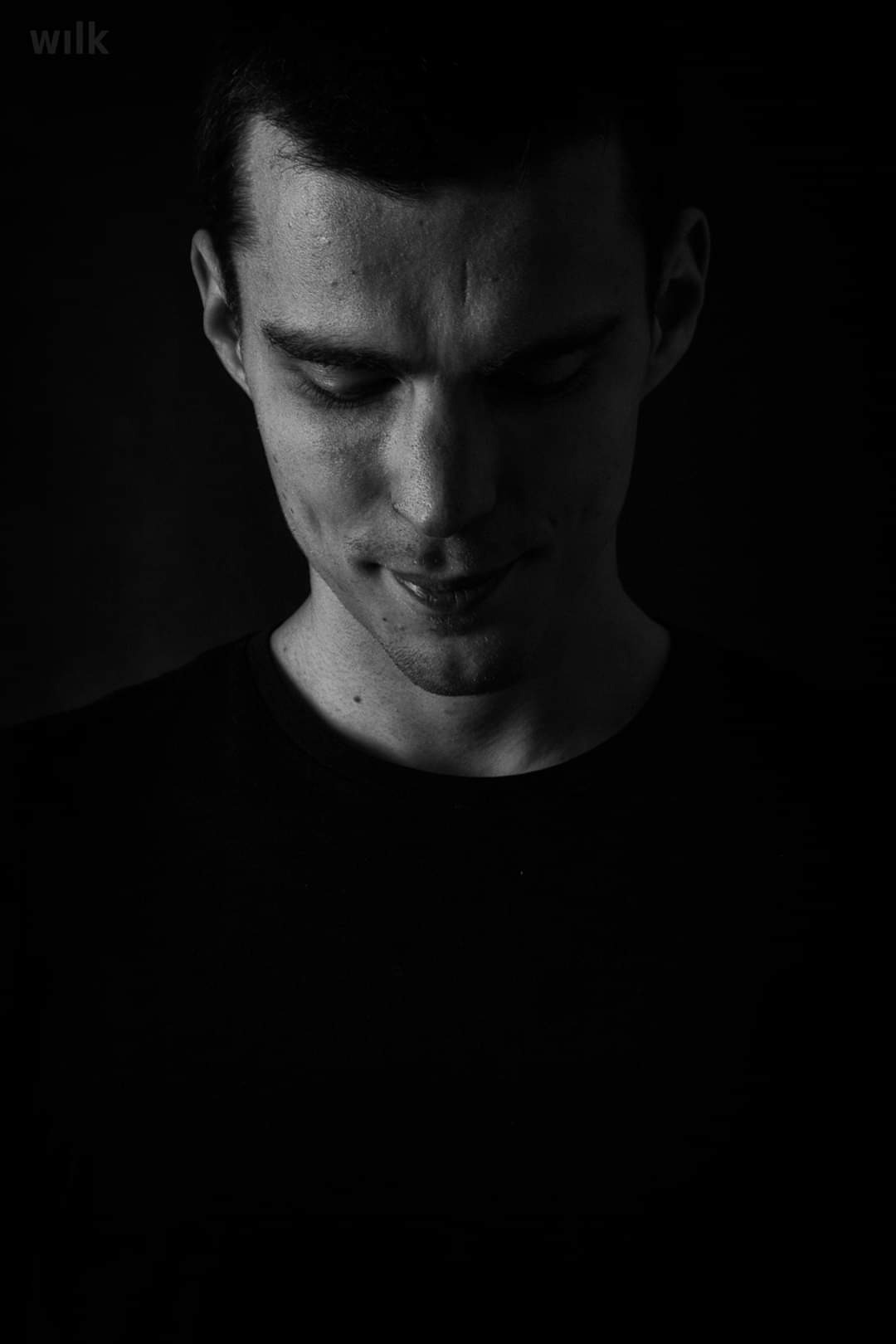 Czarno-biały portret młodego mężczyzny z lekkim uśmiechem, spoglądającego w dół, w ciemnym otoczeniu.