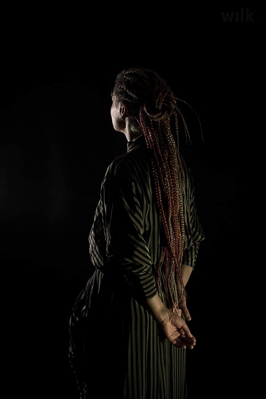 Kobieta odwrócona tyłem, z długimi, kolorowymi warkoczami, ubrana w sukienkę w pionowe paski, ręce splecione za plecami, oświetlona z boku na ciemnym tle.