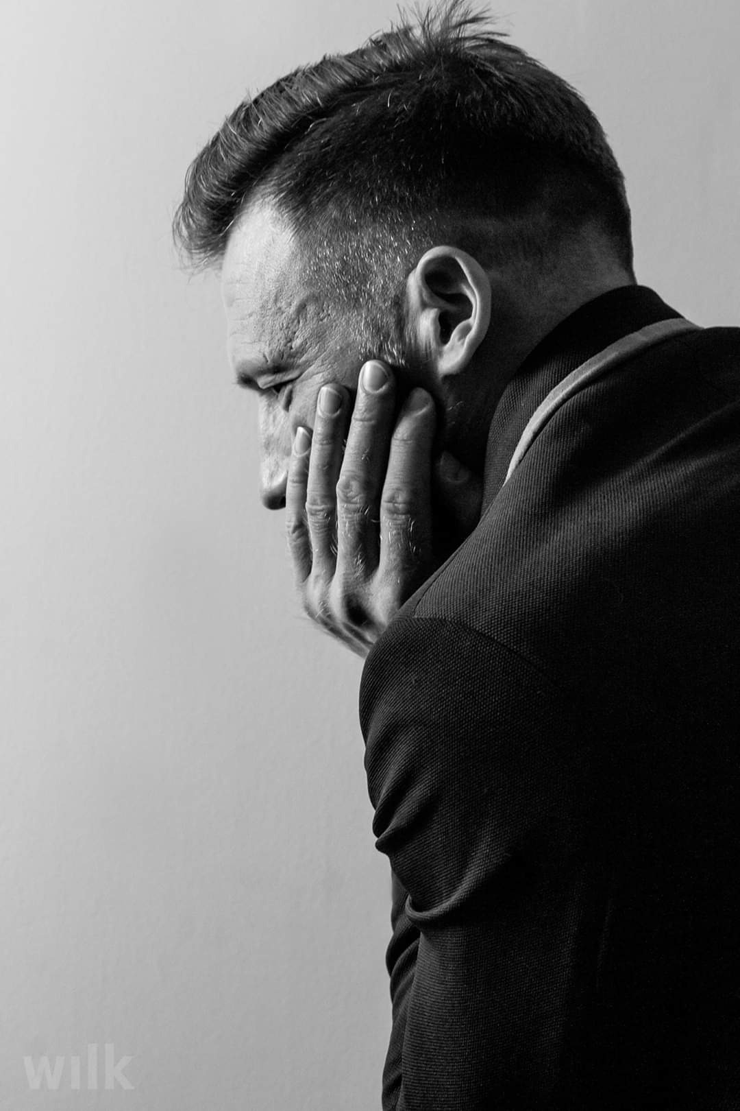 Czarno-białe zbliżenie profilu mężczyzny w garniturze, zamyślonego, z dłonią na twarzy.