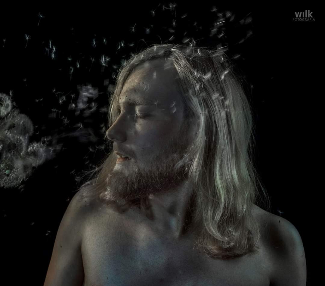 Portret mężczyzny z długimi blond włosami i brodą, dmuchającego w dmuchawiec na czarnym tle, z nasionami unoszącymi się wokół jego głowy.