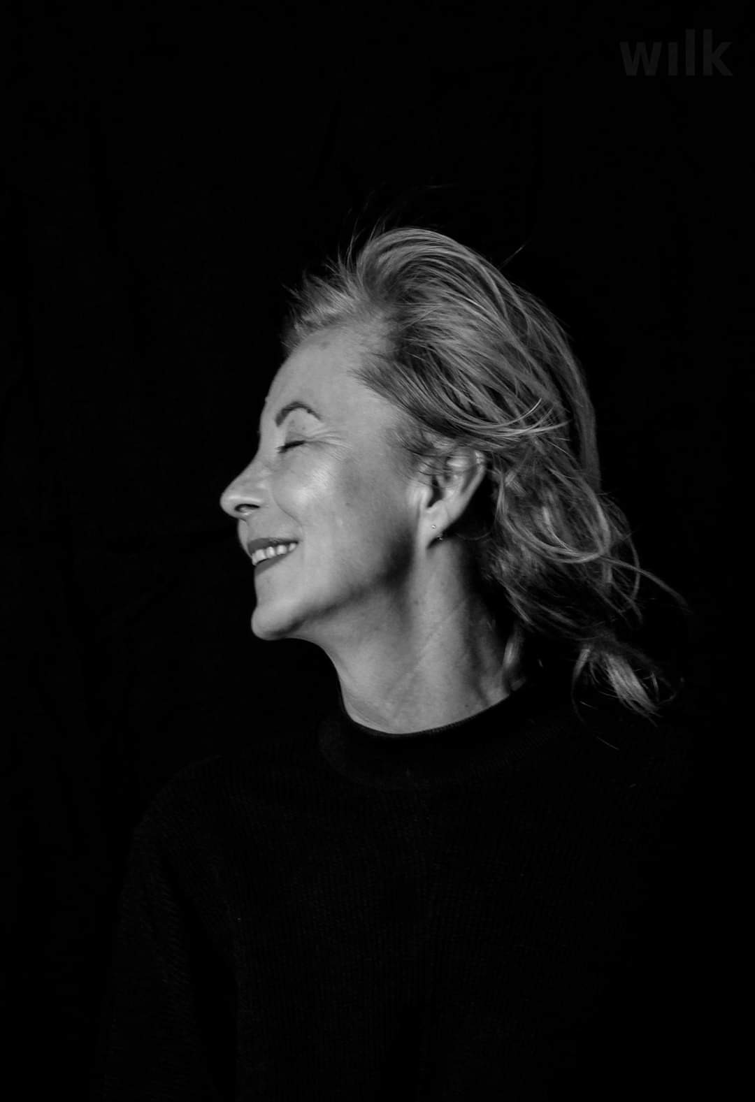 Czarno-białe zdjęcie profilowe uśmiechniętej kobiety z zamkniętymi oczami, z rozwianymi włosami na czarnym tle. Widoczny subtelny kolczyk w uchu.