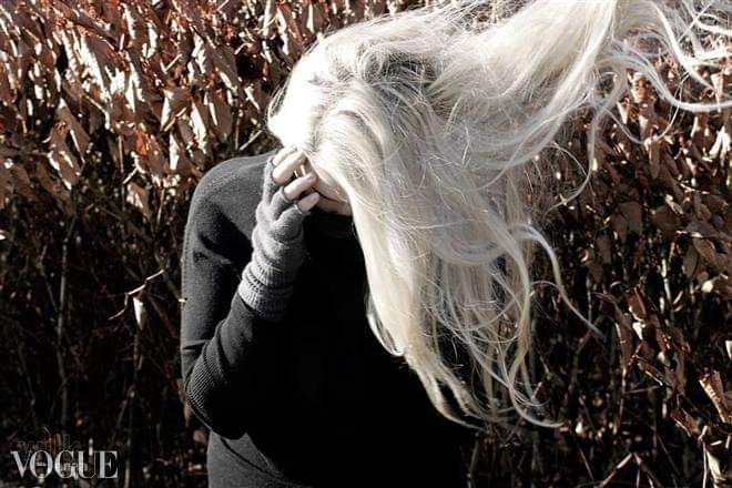 Kobieta w czarnym swetrze i rękawiczkach z odsłoniętymi palcami, z długimi, rozwianymi, siwymi włosami na tle krzewów.