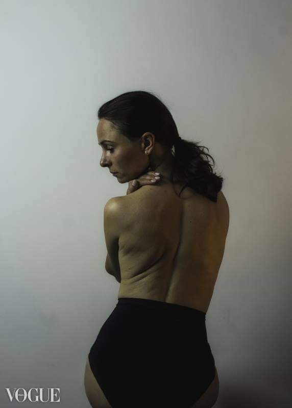 Kobieta z ciemnymi włosami spiętymi w kucyk, pozuje tyłem, z rękami skrzyżowanymi na szyi, ubrana w czarne figi na tle jasnej ściany. Widoczny znak wodny 'Vogue'.