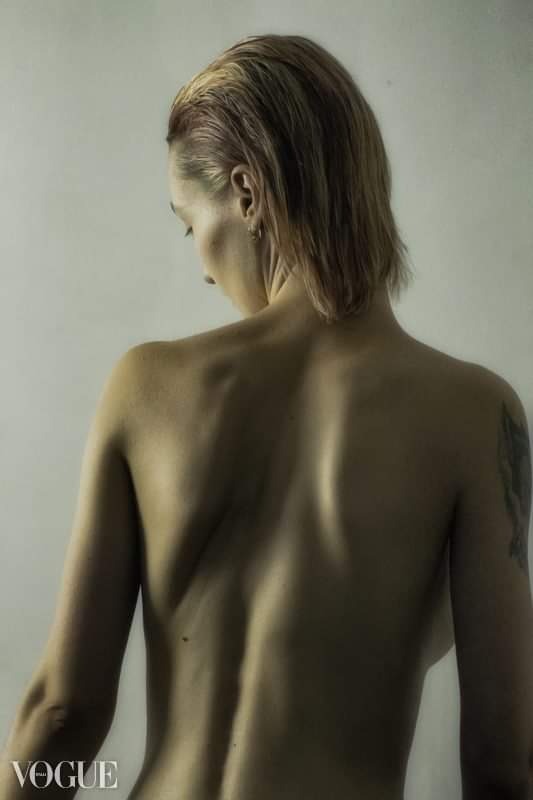 Kobieta z krótkimi blond włosami, widok z tyłu, odsłonięte plecy z widocznym tatuażem na ramieniu, delikatne oświetlenie, minimalistyczne tło, logo VOGUE w lewym dolnym rogu.