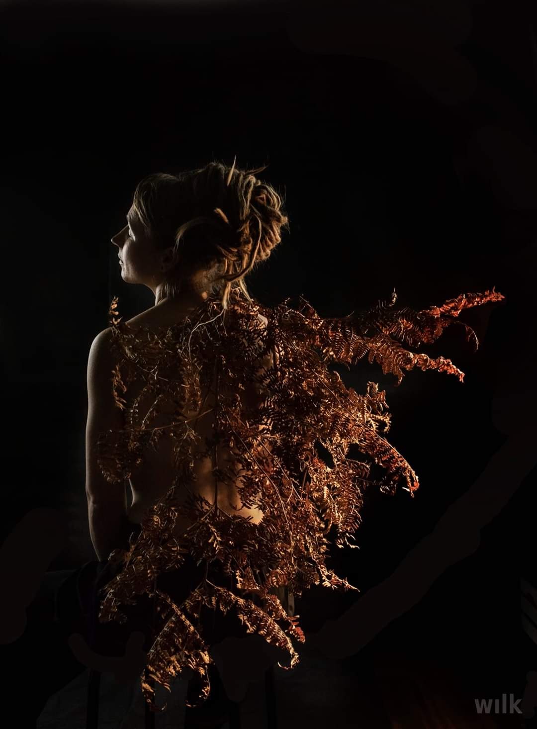 Kobieta z profilu, z upiętymi włosami, na plecach kompozycja z suszonych paproci w ciepłych odcieniach brązu, na czarnym tle.