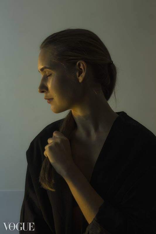 Portret kobiety z profilu, oświetlona ciepłym światłem, trzymająca w dłoni kosmyk włosów, na jasnym tle, w dolnym rogu widoczne logo magazynu Vogue.