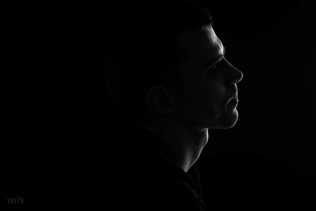 Czarno-biały portret profilowy mężczyzny, oświetlonego z boku, z wyraźnie zarysowanymi konturami twarzy na ciemnym tle.