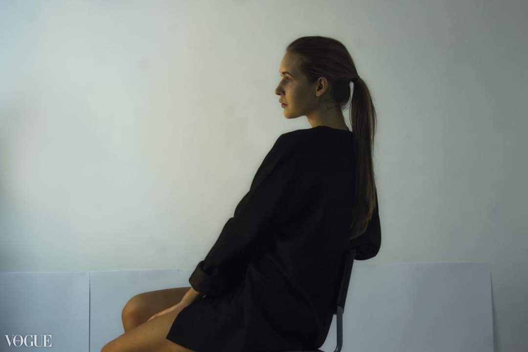 Kobieta siedząca bokiem na krześle, ubrana w czarną oversize'ową marynarkę, z włosami spiętymi w kucyk, na białym tle.