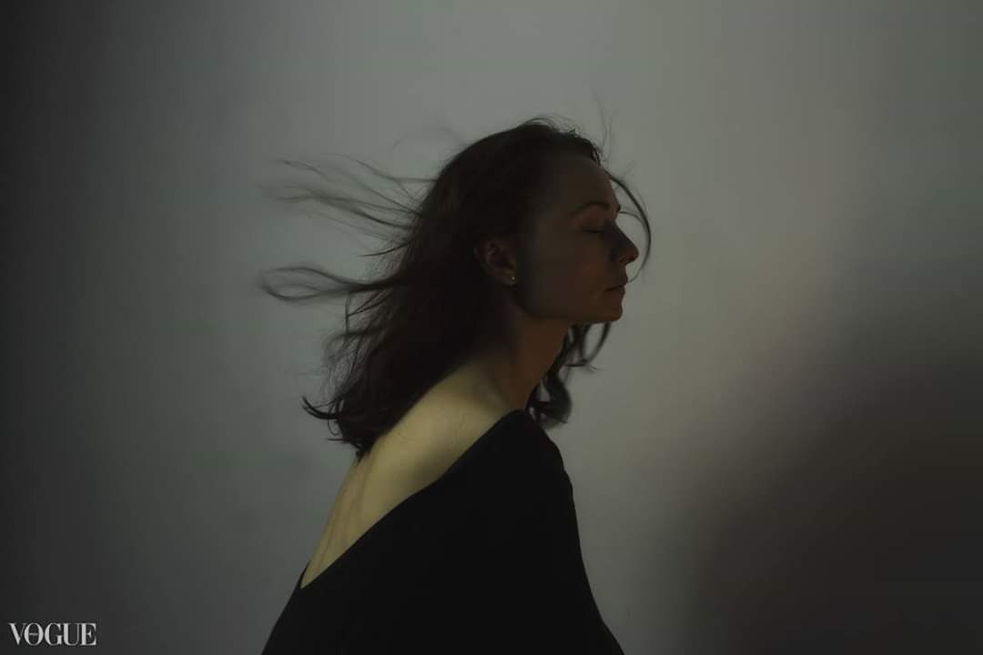 Portret kobiety z zamkniętymi oczami, profil, z rozwianymi włosami na szarym tle, w czarnej sukience z odkrytymi plecami, dolny lewy róg zajmuje logo Vogue.