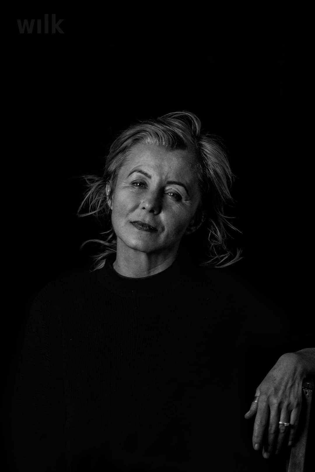 Portret kobiety w czerni na ciemnym tle, z subtelnym uśmiechem, widocznym kolczykiem w nosie i pierścionkami na dłoni opierającej się o coś niewidocznego.